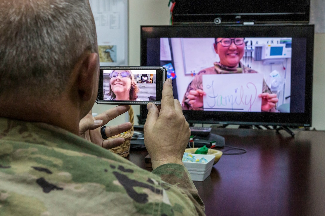 A soldier holds up a phone that has his wife on screen, while looking at a person on another screen who holds a sign that says "Family"