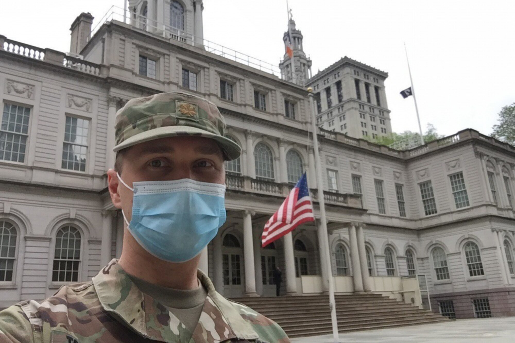 A man dressed in a military uniform and wearing a face mask poses for a photo in front of a large building where the flag is flying at half-staff.
