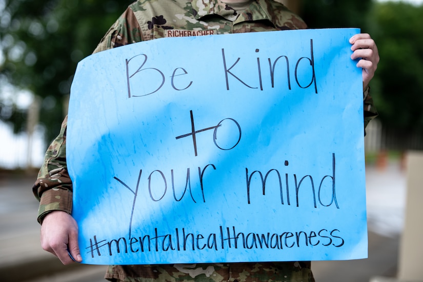 An airman holds up a handmade blue sign reading "Be kind to your mind" with a "#mentalhealthawareness" hashtag.