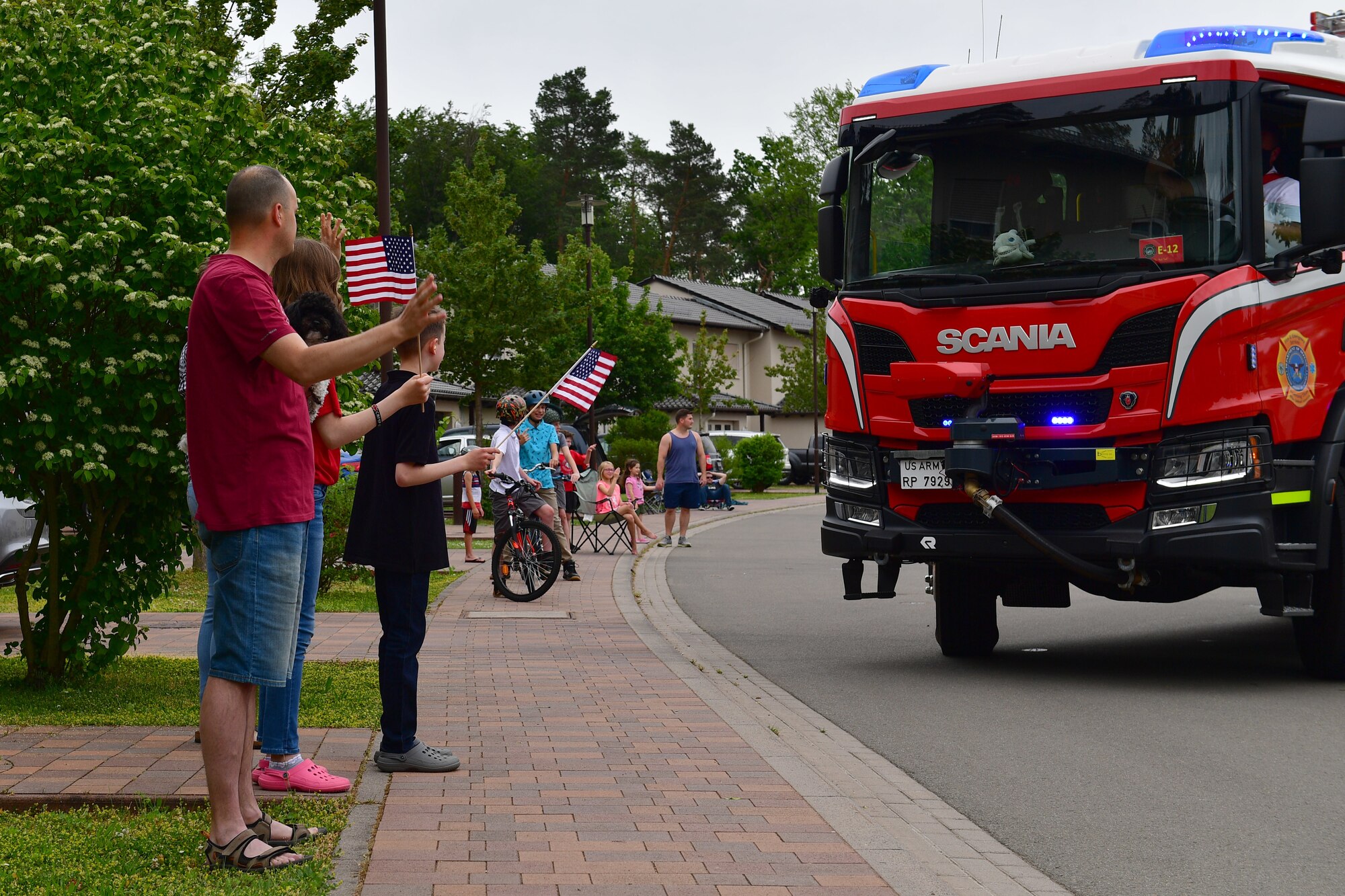 Military families stand on a sidewalk and wave to a passing fire truck.