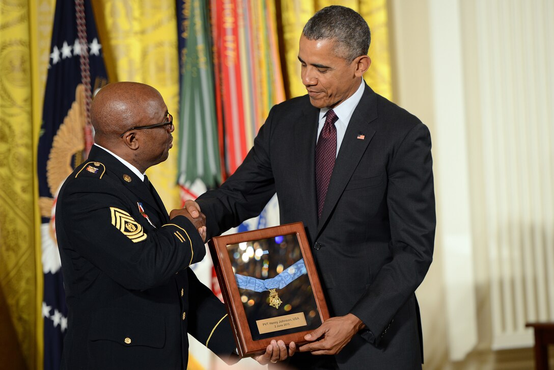 A man shakes hands with another man. Both hold a plaque.