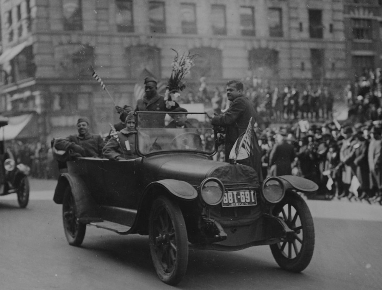 Two men stand and four others sit in a car as they wave to a crowd during a parade. One of the men who is standing is holding a bouquet of flowers.