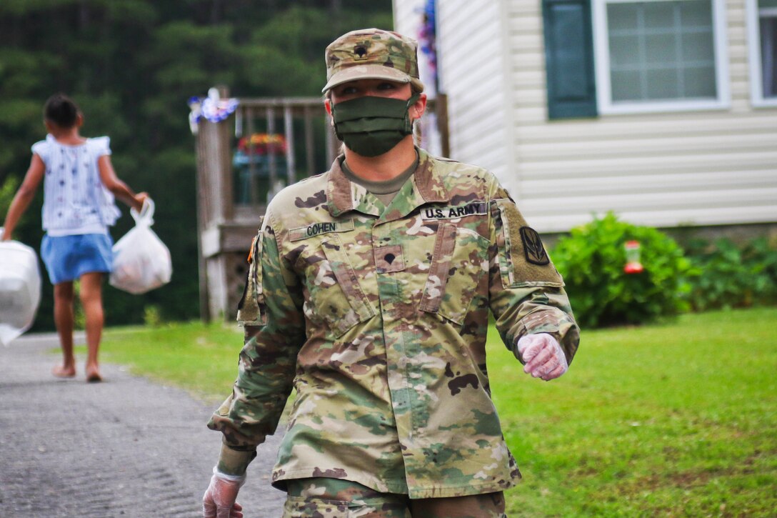 A soldier wearing a face mask walks down a driveway as a resident walks in the opposite direction carrying a bag in each hand.