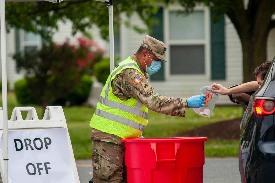 A soldier wearing personal protective equipment takes a plastic bag from a person in a car.