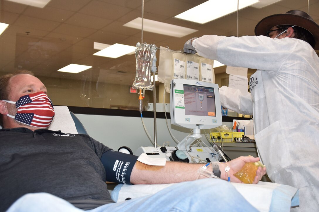 A man wearing an American flag face mask sits in a donation chair while a medical professional adjusts a machine above his head.