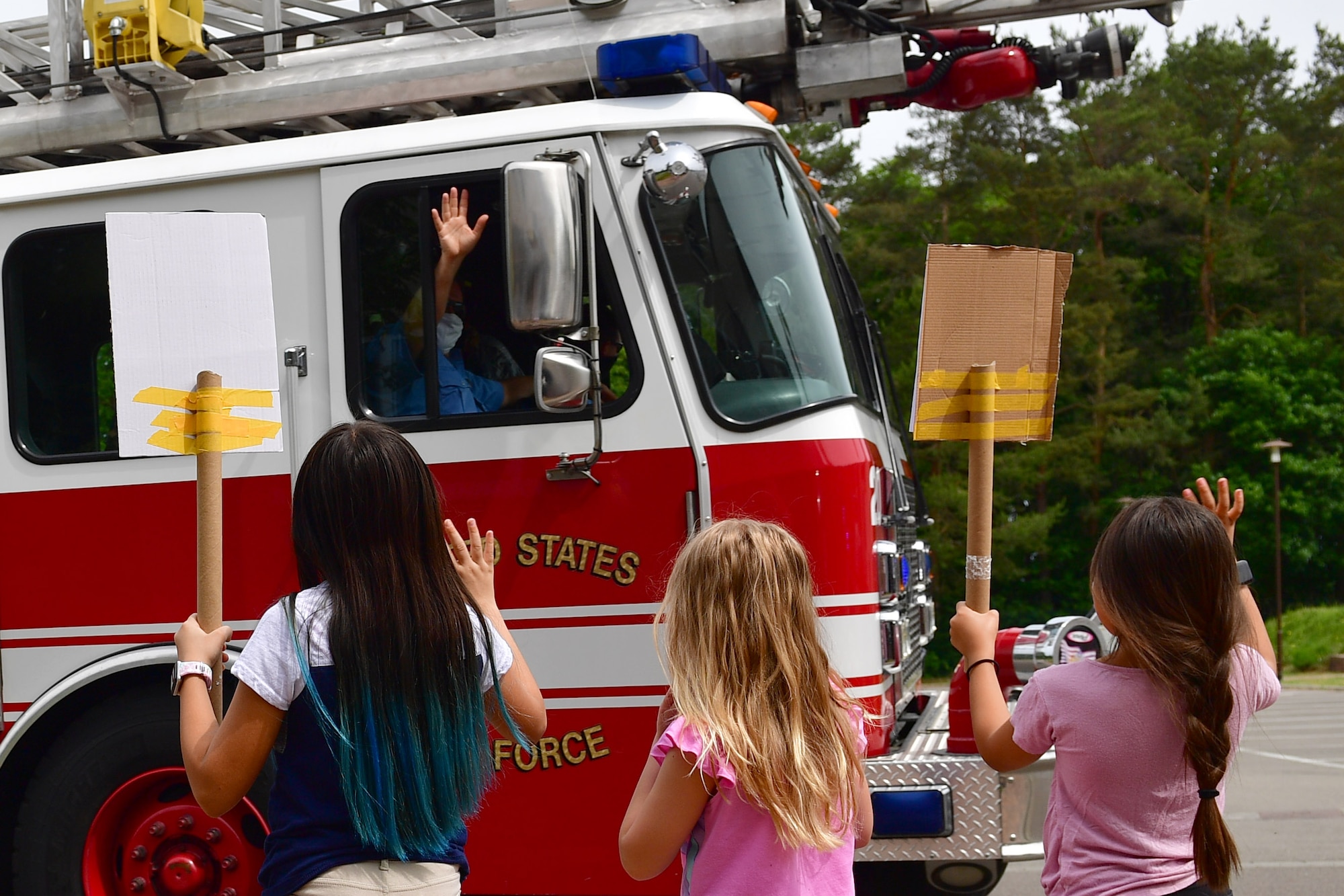 Three children hold signs and wave to first responders in a passing fire truck.