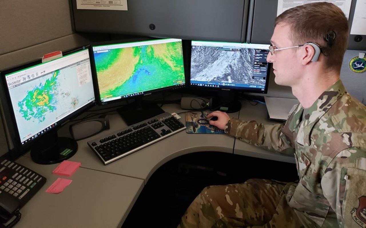 An airman operates a computer with three weather-related screens.