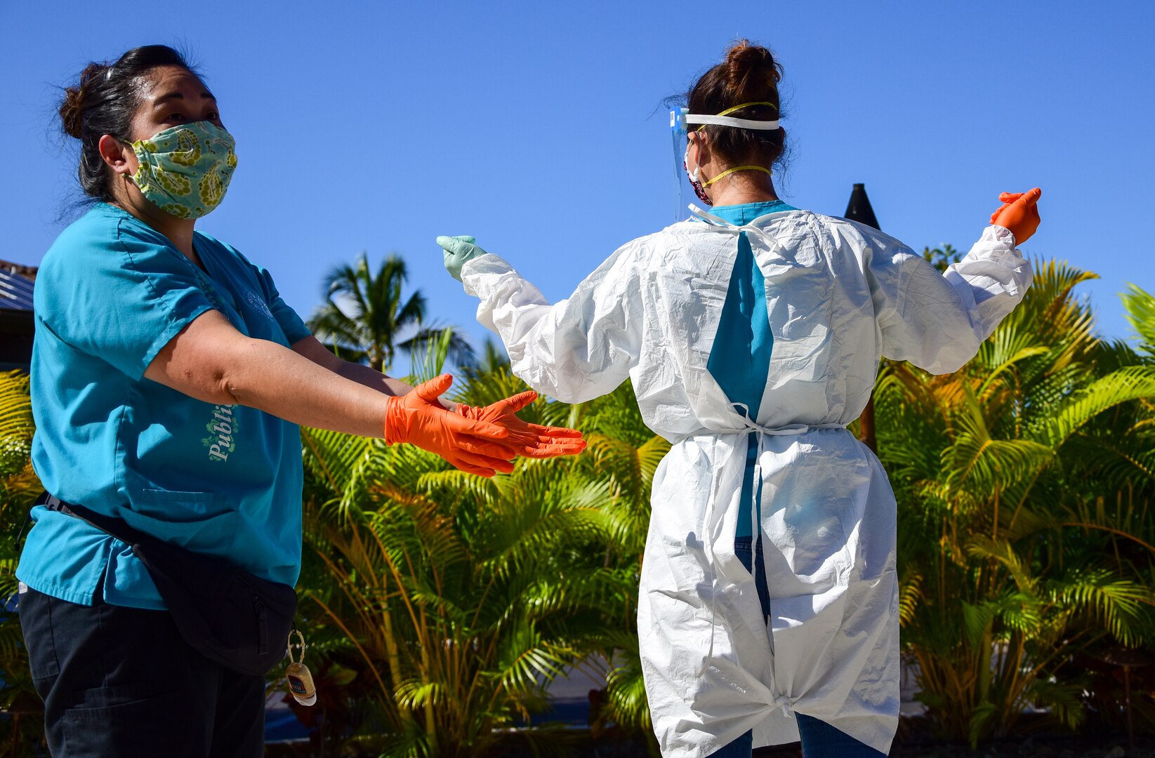 One woman wearing orange protective gloves stretches her arms toward another woman wearing a white personal protective gown outdoors.