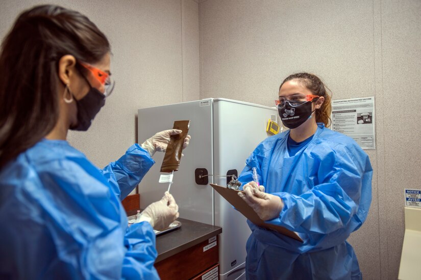 A woman wearing personal protective equipment documents and verifies if a patient has received a vaccine or placebo. Another woman holds a clipboard.