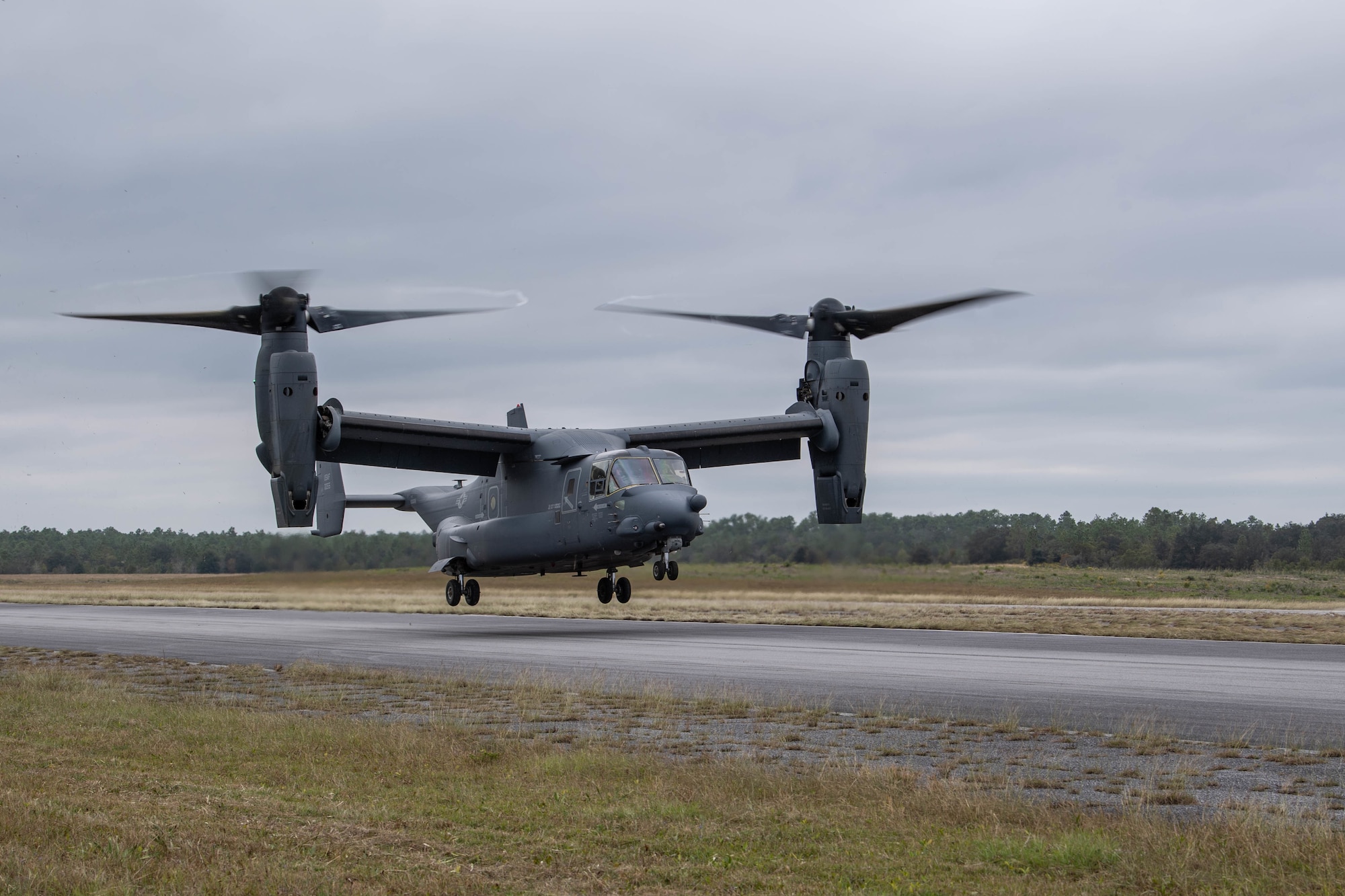 A dual-rotor helicopter is just about to land on a road in a field. Its rear wheels are close to touching the road as it lands tail-first a few hundred feet away on a road that we are standing beside.