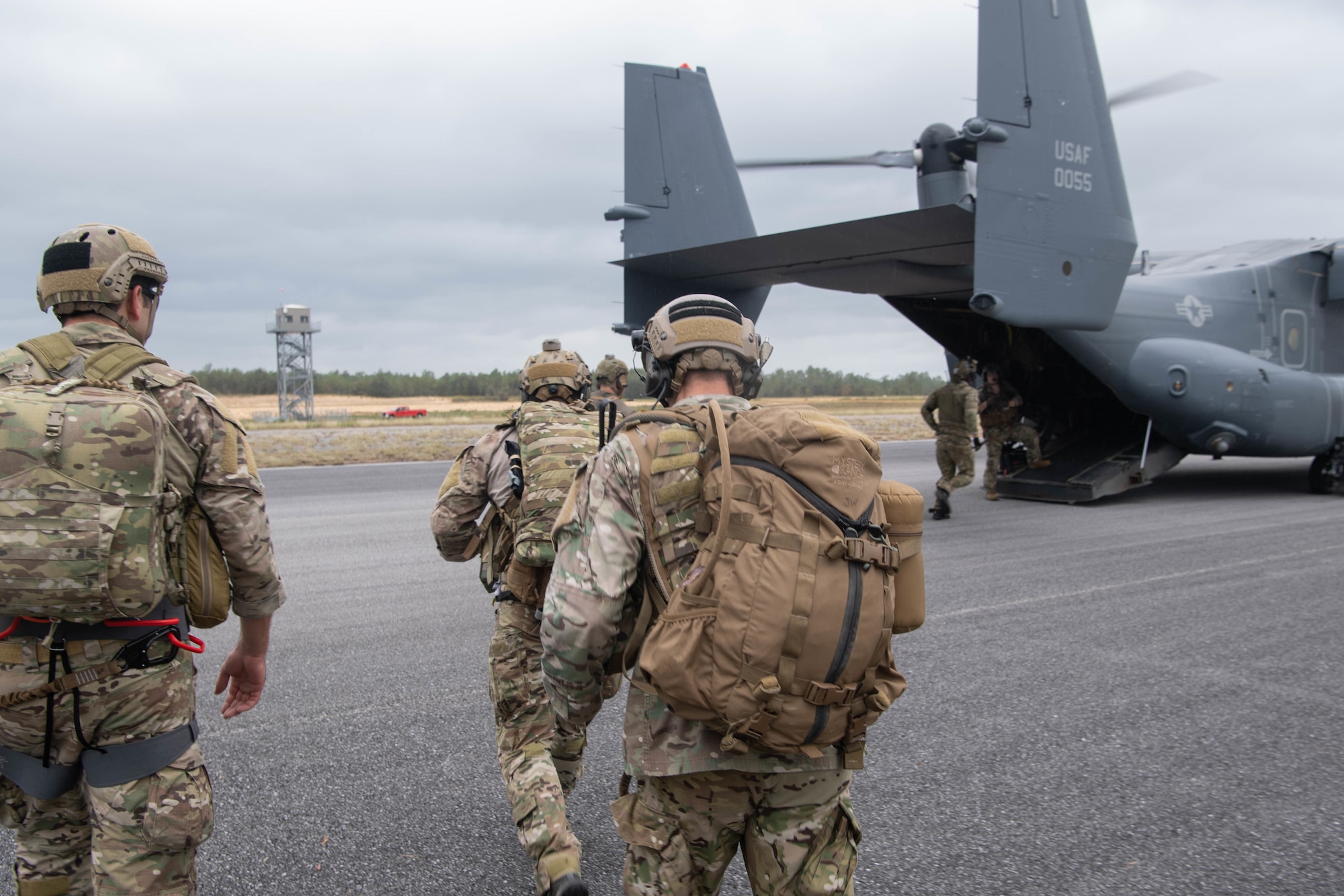 A trio of Special Tactics operators in the foreground with heavy packs walk toward a waiting dual-rotor helicopter ramp where another operator is approaching the ramp.