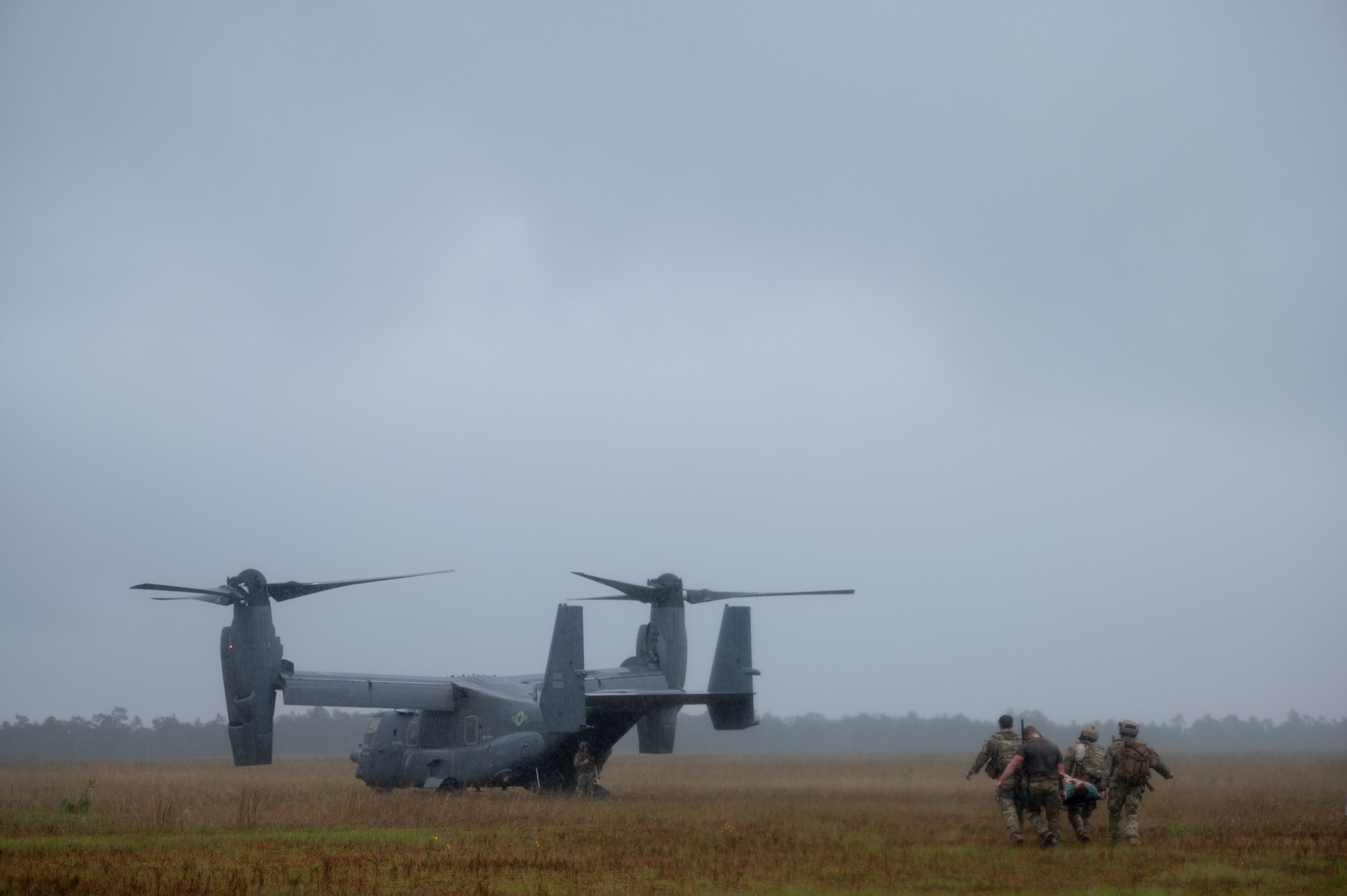 Four Special Tactics Operators carry a patient on a stretcher to a waiting dual-rotor helicopter waiting in a field.