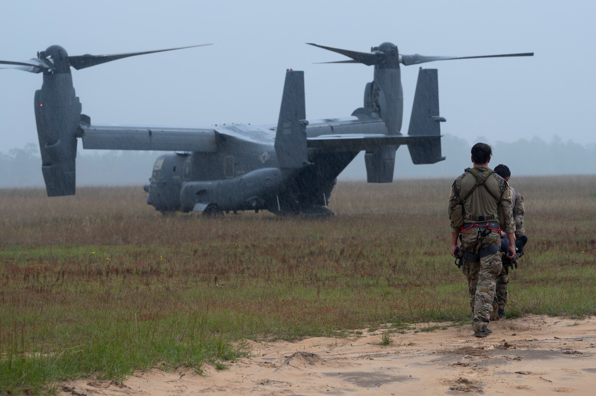 Two Special Tactics Operators carry a patient on a stretcher to a waiting dual-rotor helicopter waiting in a field.