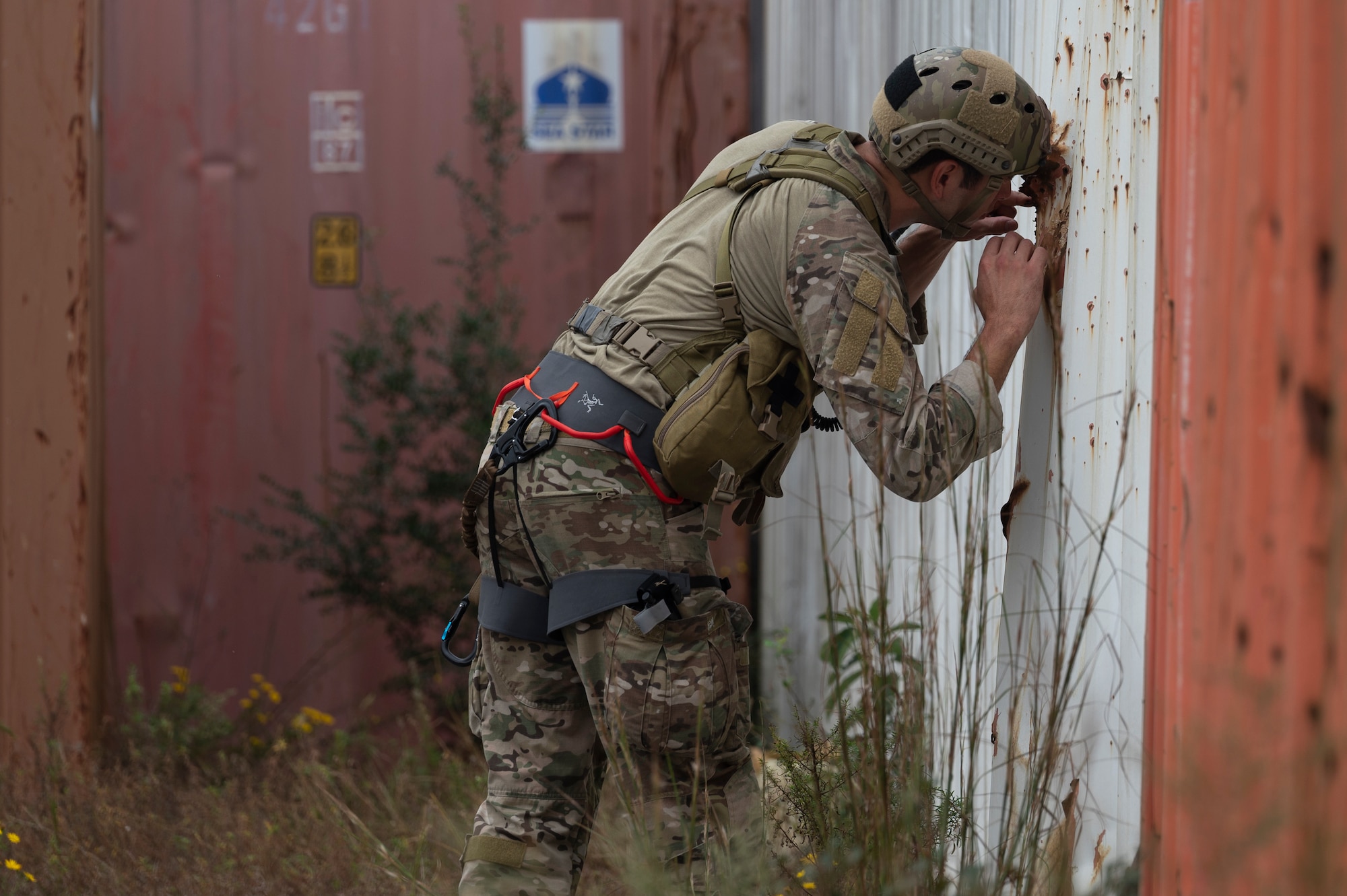 We look at the side of a Special Tactics Operator as he leans over with his helmet against a corrugated metal wall as he looks for injured civilians through a hole in the wall.
