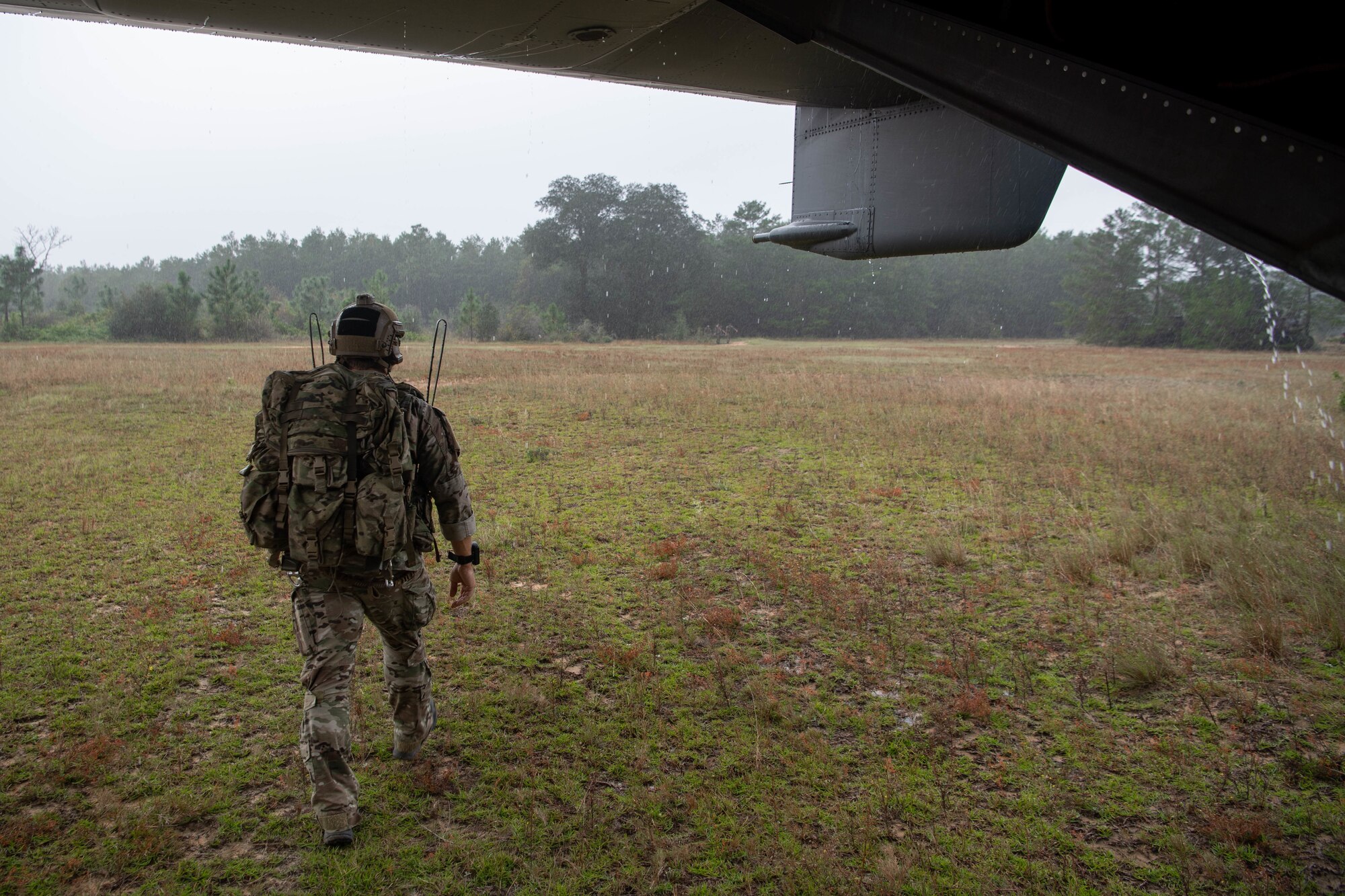 A single Special Tactics Operator with heavy pack walks away from us under the tail of a helicopter towards a simulated disaster site in the tree line in the distance.