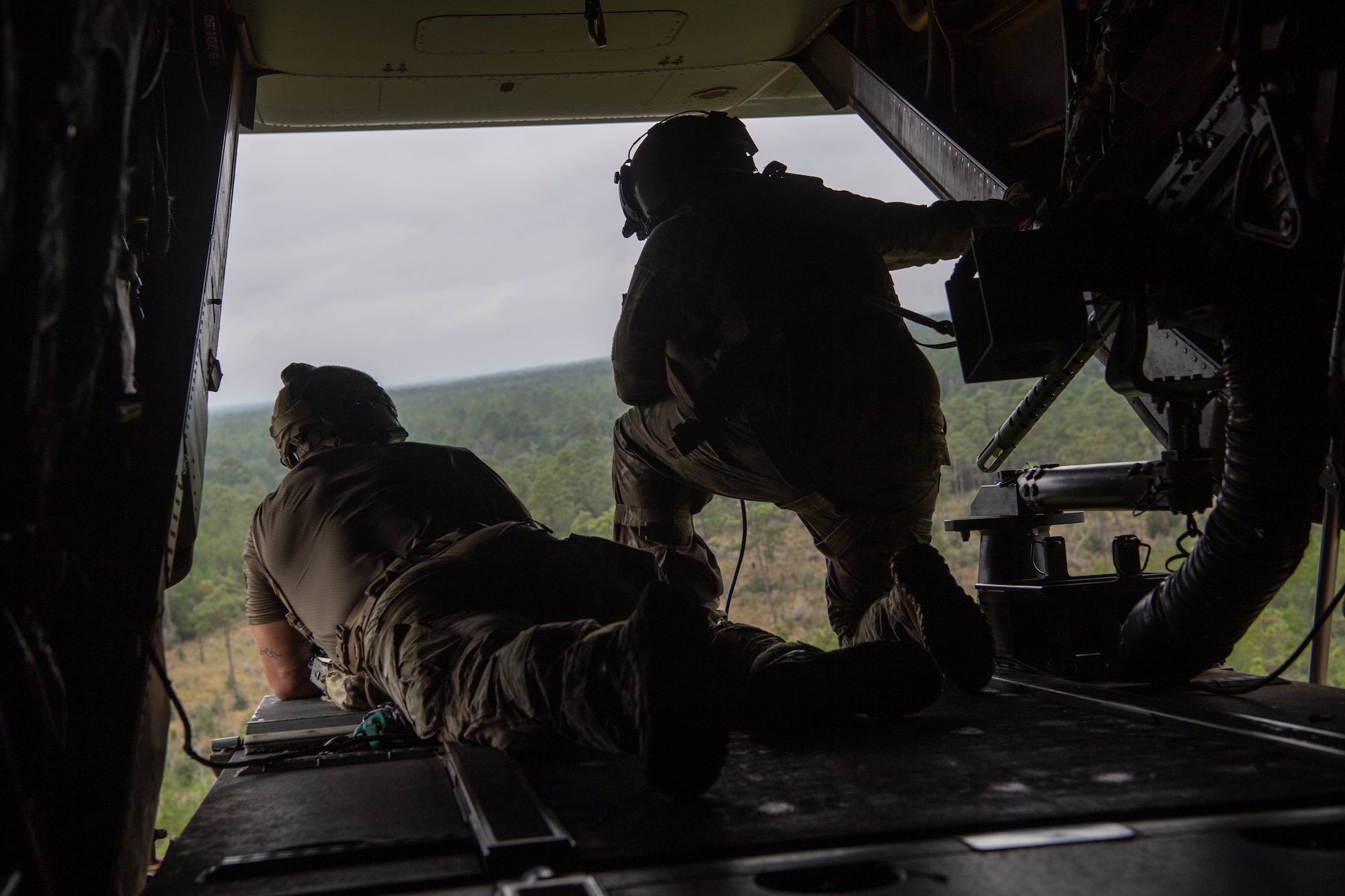 As we look out of the open ramp of a dual-rotor helicoper, a Special Tactics operator lies on the left as a kneeling flight engineer operates equipment to his right as they survey the ground below the helicopter looking for victims of the simulated disaster.