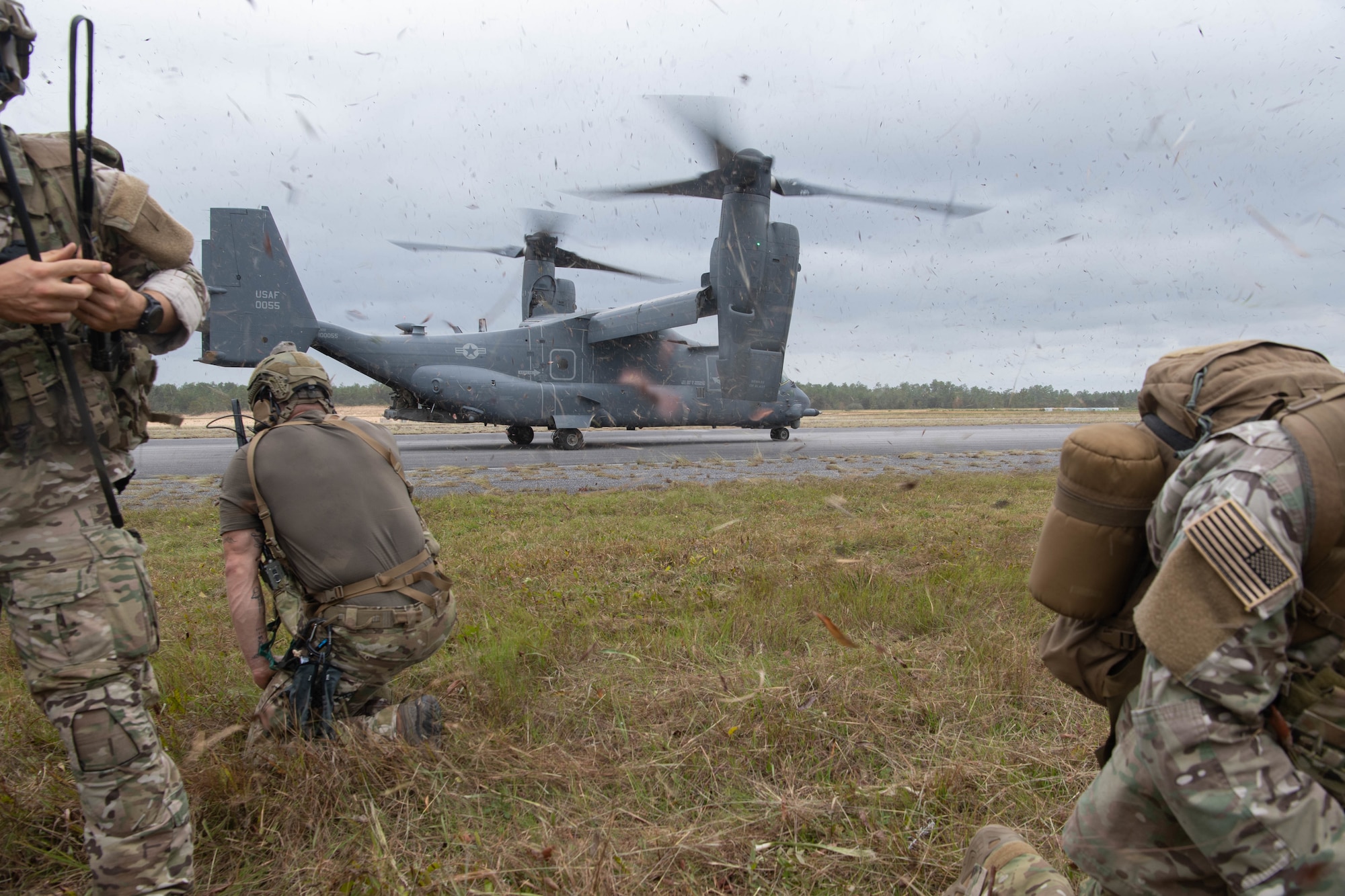 A trio of Special Tactics operators in the foreground brace as a dual-rotor helicopter a 100 feet away spins up its rotors prior to take-off.