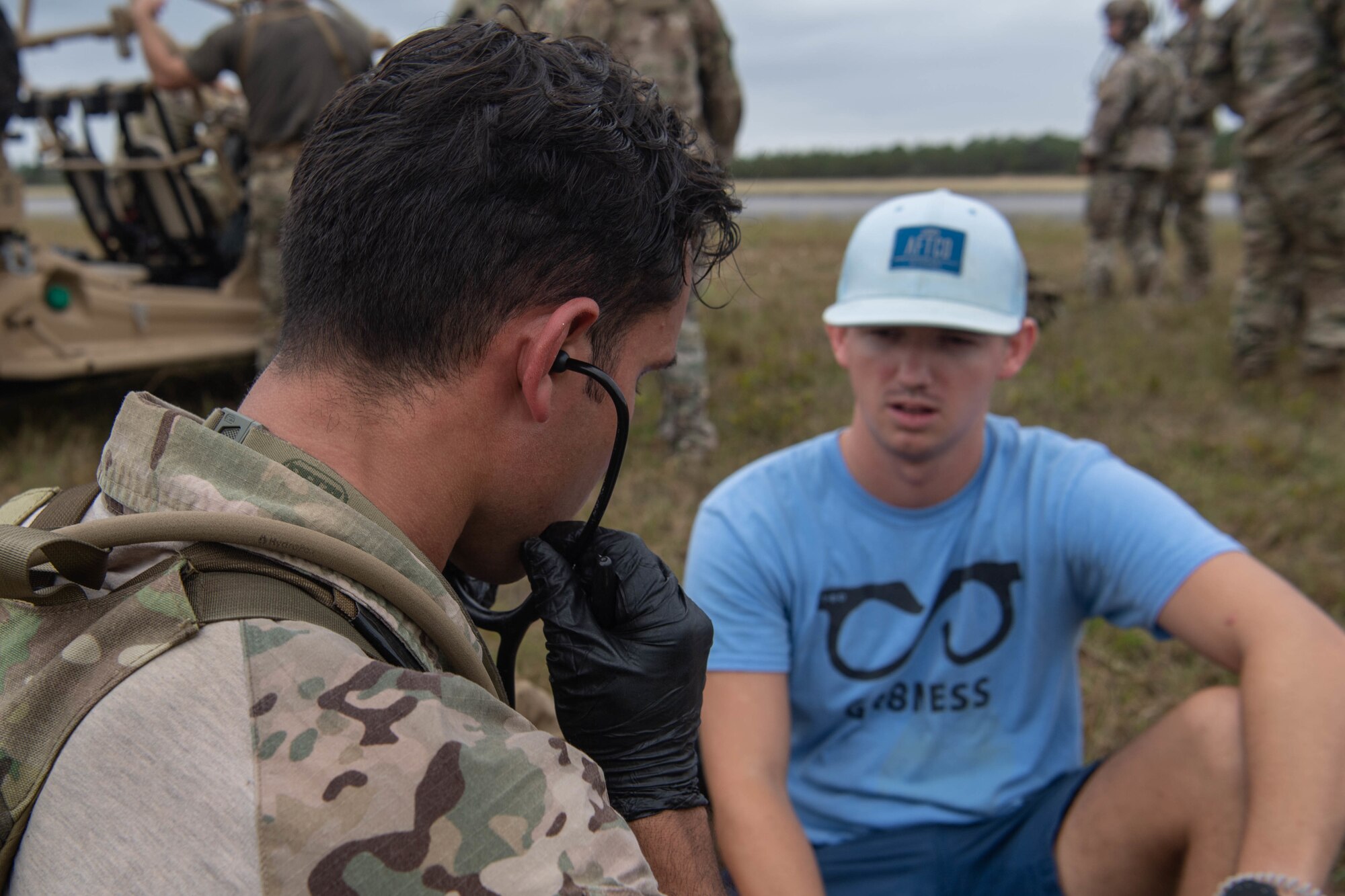 We look over the shoulder of a Special Tactics operator as he inserts a stethoscope into his ears so he can listen to a patient sitting on the ground to his right in a blue t-shirt and black shorts