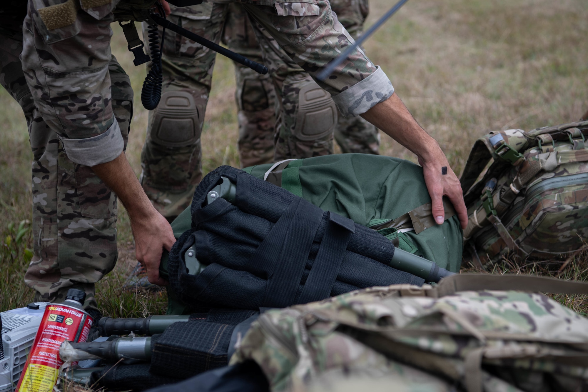 We see the arms of a Special Tactics operator on the left as he reaches down to pick up a folded-up dark green stretcher from a pile of equipment. We see the legs of another operator as he stands behind the operator reaching for the stretcher.
