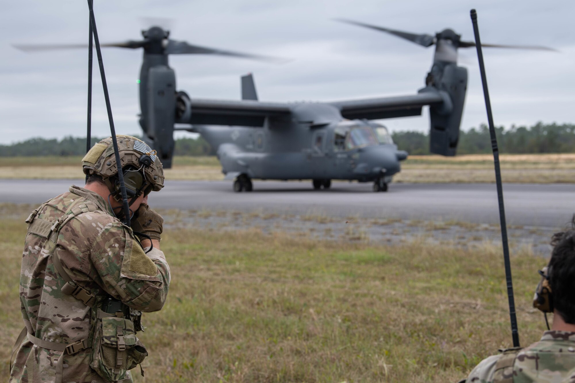 Two Special Tactics operators wearing backpacks with tall antennas stand near us beside a road where a dual-rotor helicopter has landed. The operators are using their radios to talk to the pilot of the helicopter.