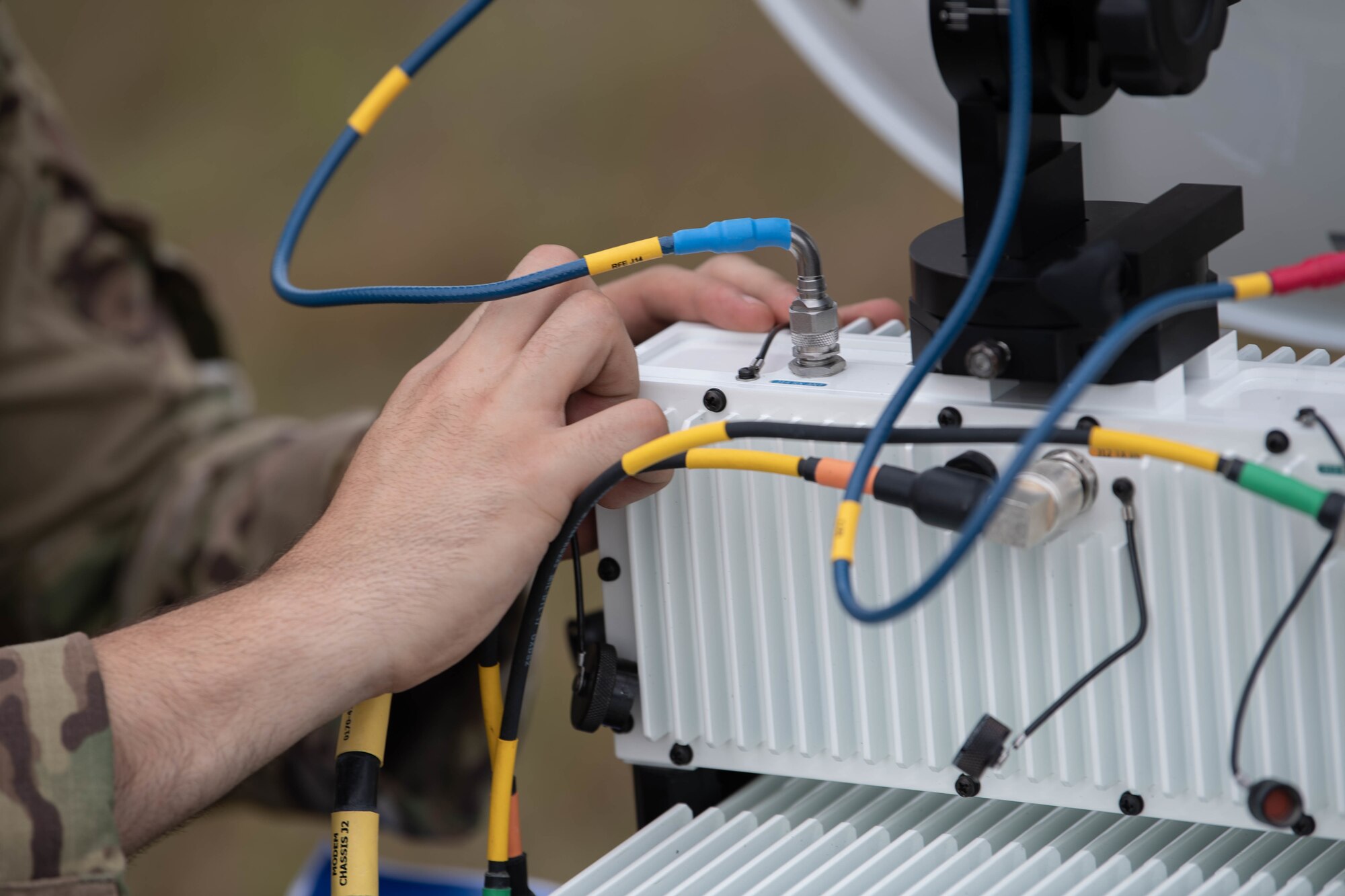 We see a stack of white boxes with black wires attached to them. We see the technician's hands as he works on the white boxes, connecting the black wires with brightly colored labels to the boxes so that Special Tactics operators can use them for communications during an emergency.