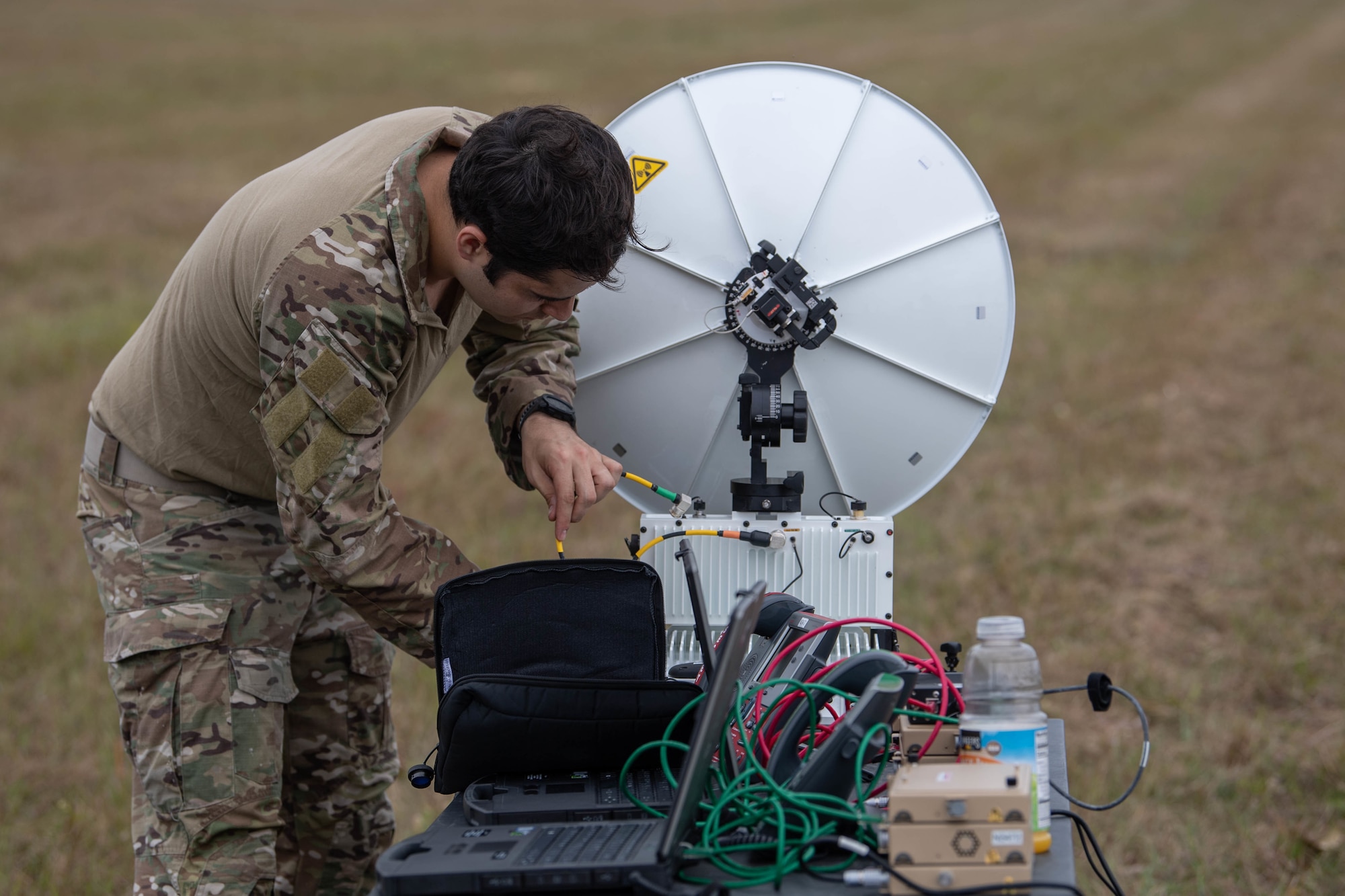 We are looking down a table at a small satellite dish pointing away from us. A Special Tactics operator is connecting radio equipment to the dish to aid communications during the disaster response exercise.