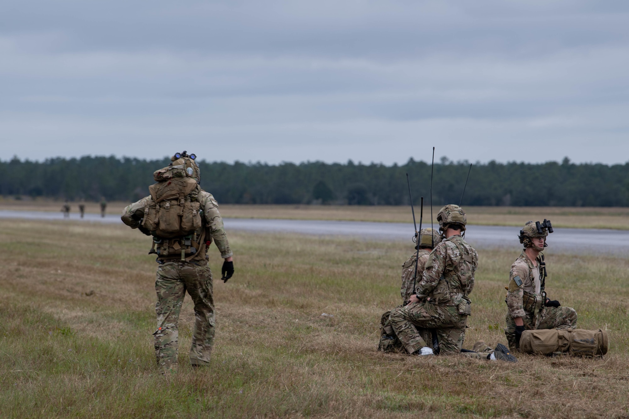 A trio of Special Tactics operators gather. One is walking in from the left, a radio operator is kneeling beside his gear in the middle of the picture, and another operator is sitting on the ground to the right.  Off in the far distance another trio of operators can be seen standing in a line of three across the road.