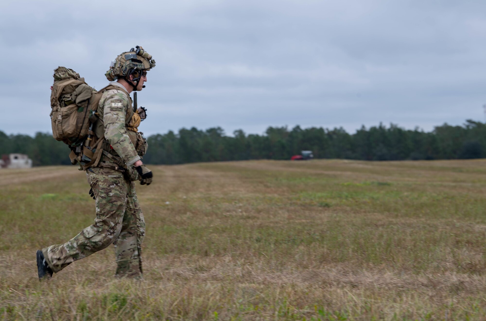 A Special Tactics operator walks close to us from from left to right across a field with a heavy pack. We see a large field of grass stretching off into the distance where it meets a tree line.
