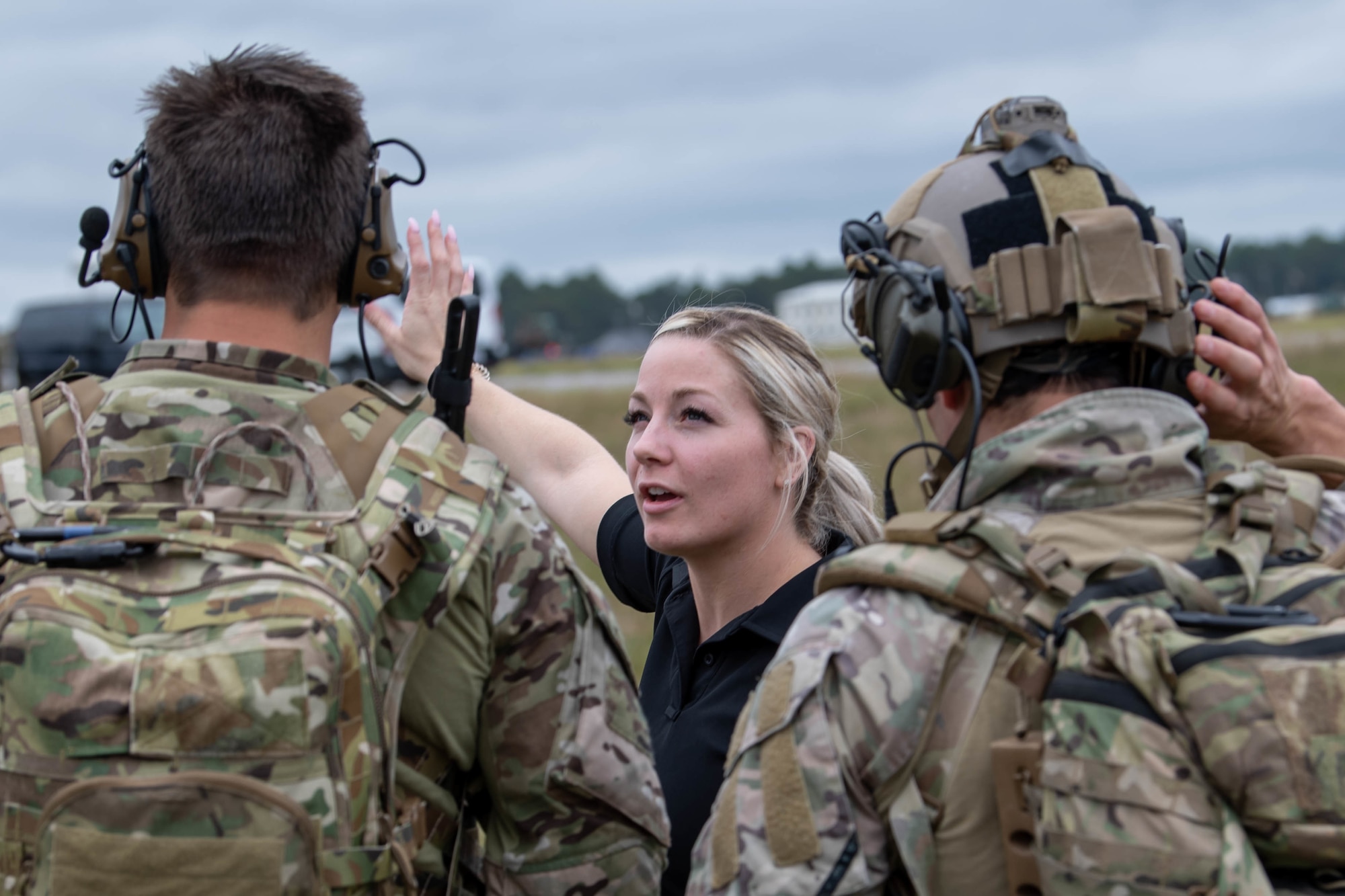 A blond woman in a black t-shirt can be seen talking to a pair of Special Tactics operators as we look between them over their shoulders at the woman gesturing toward the distance.