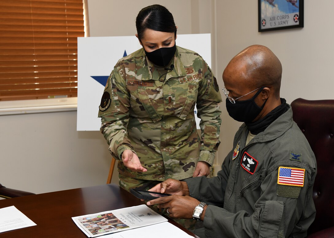 Airman that is standing talks to sitting Airman using phone.
