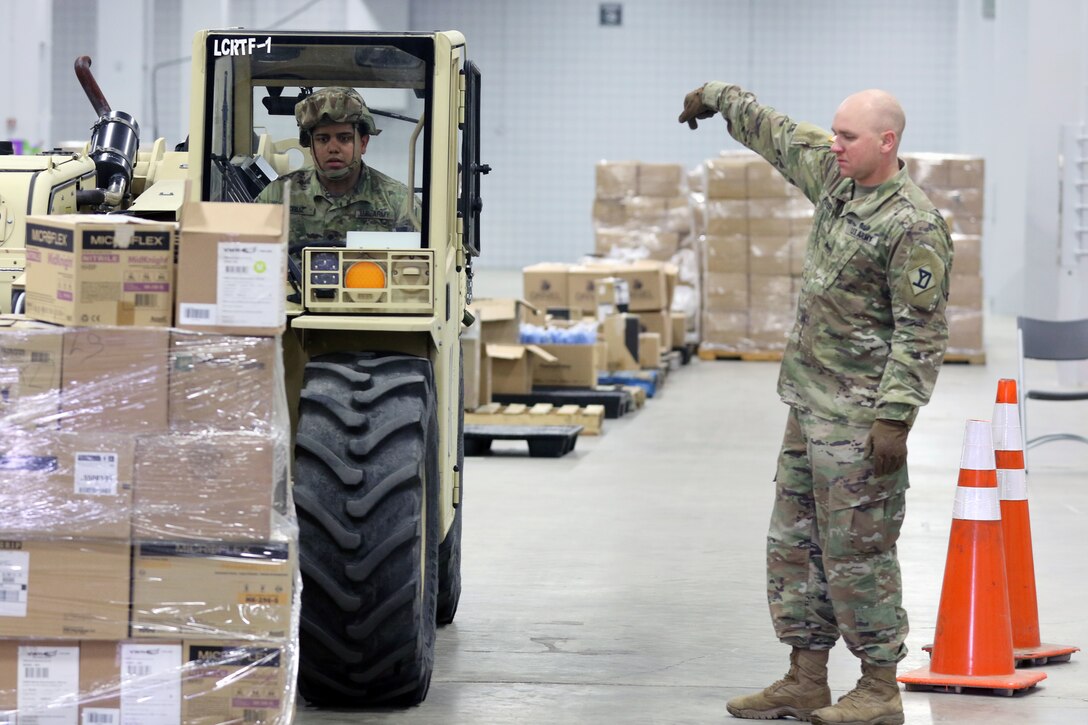 Male soldier directs another male soldier on a forklift where to place supplies in a warehouse.