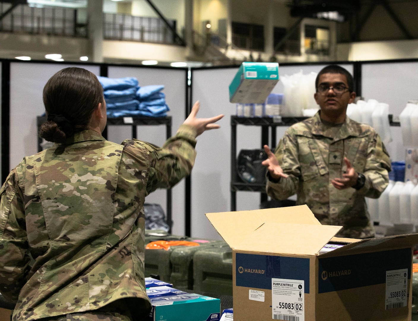 A female soldier tosses a small box in the air toward a male soldier in a storage room.