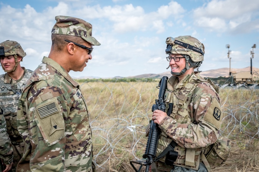 An older man in a military uniform talks to a young woman in a military uniform as they stand in a field.