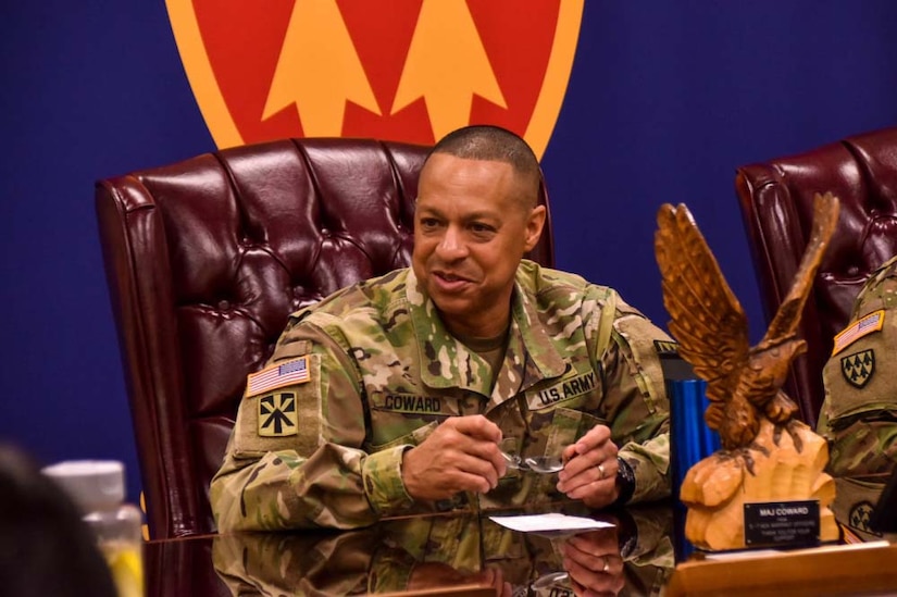 A man in a military uniform sits at a table in a large chair; he is holding a pair of glasses in his hands.