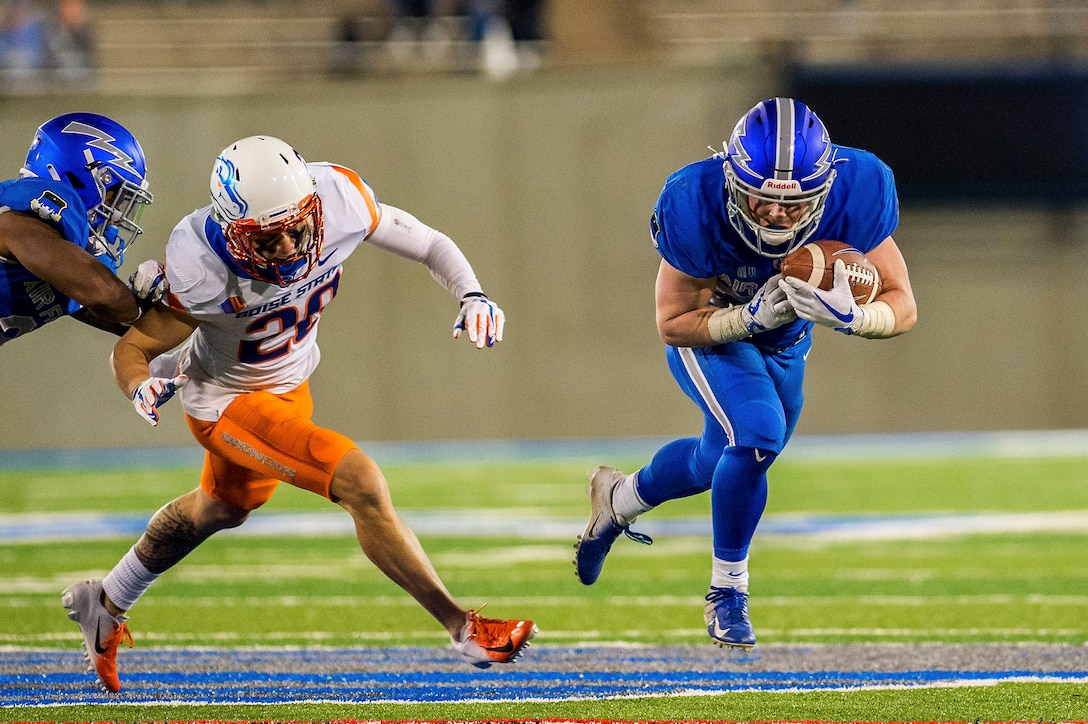 A football player runs while carrying a football as another teammate blocks another player.