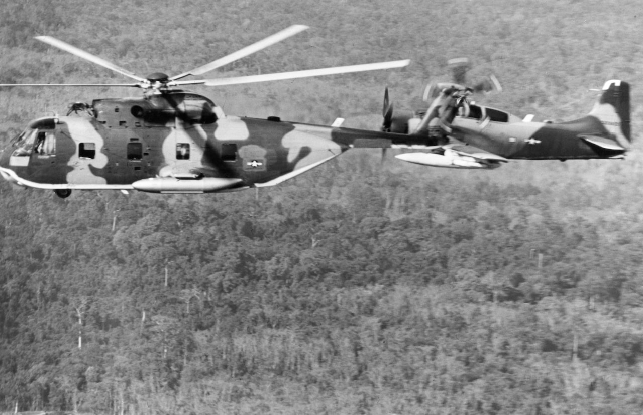 A camouflage-painted helicopter flies beside an airplane over a jungle.