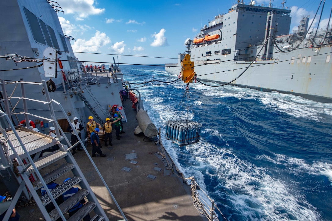 Two large ships steam next to each other as a pallet of supplies is moved from one ship to another.