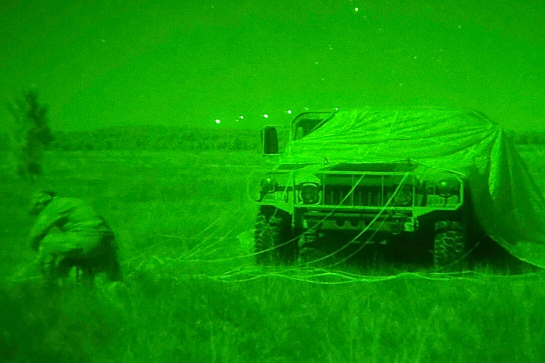 A solider illuminated by green light packs a parachute next to a military vehicle.