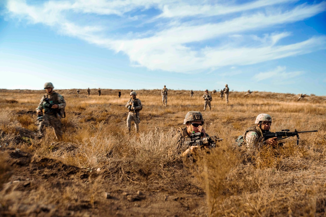 Two Marines holding weapons kneel in desert terrain as others walk behind.