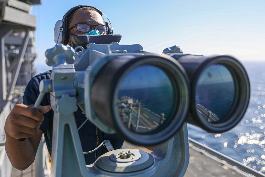 A sailor looks through large binoculars on the deck of a ship.