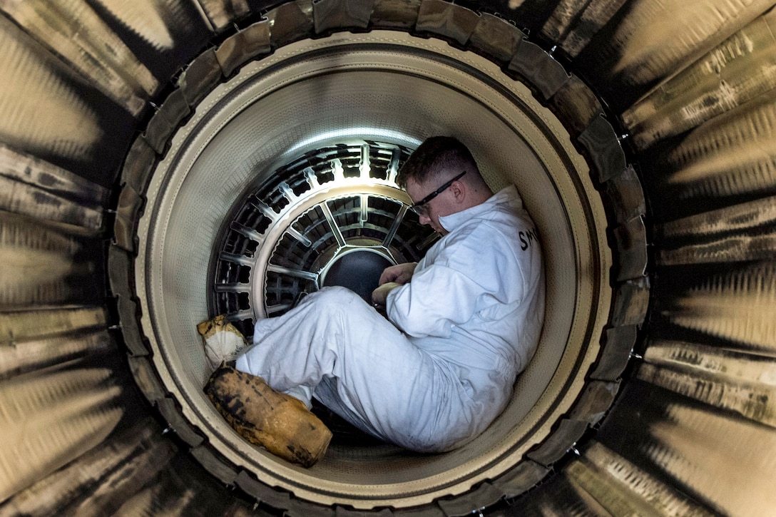An airman in white coveralls sits inside a metal circular structure.
