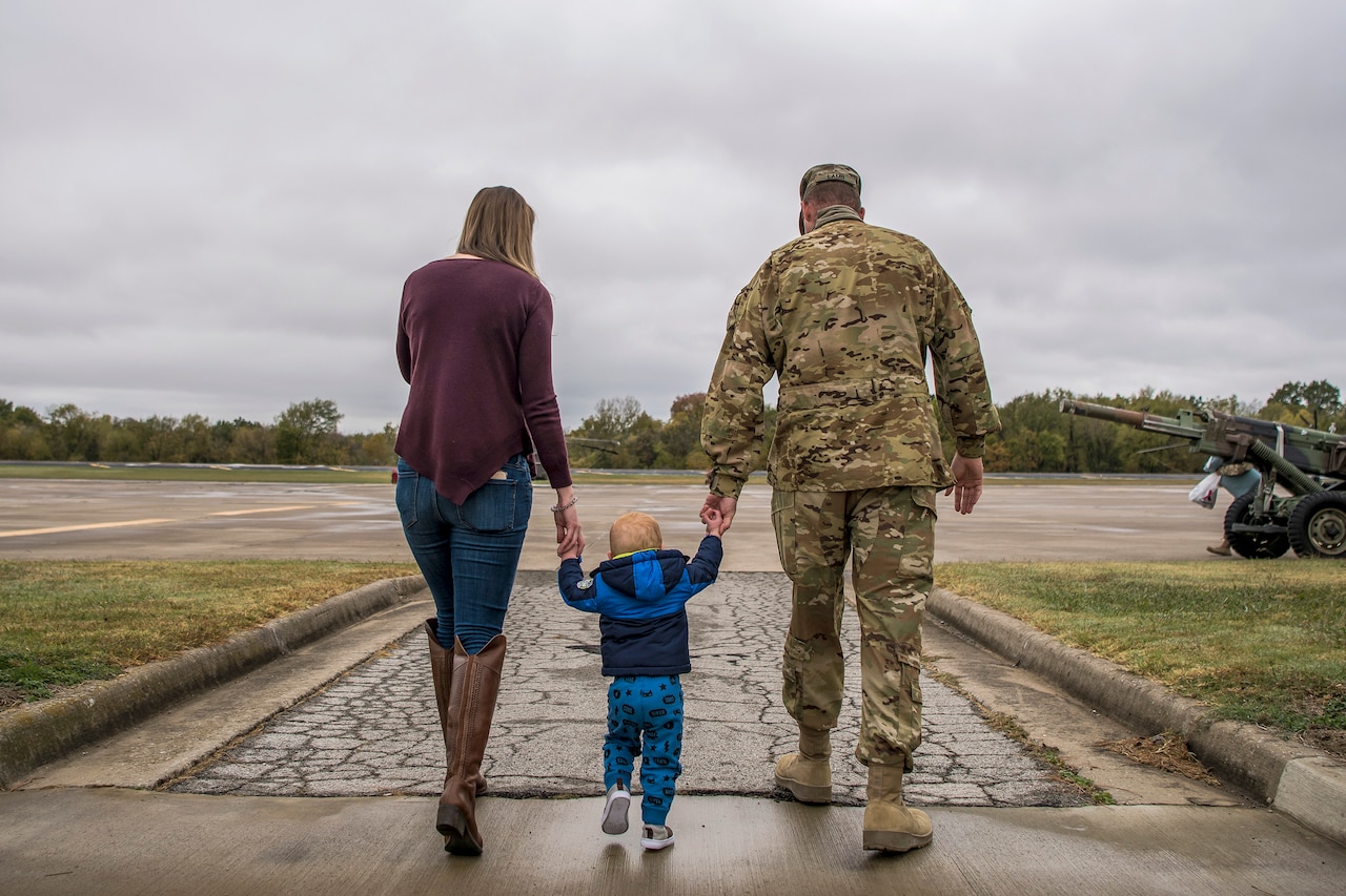 A child holds hands with an Army National Guardsman and a civilian as they all walk together on a paved path.