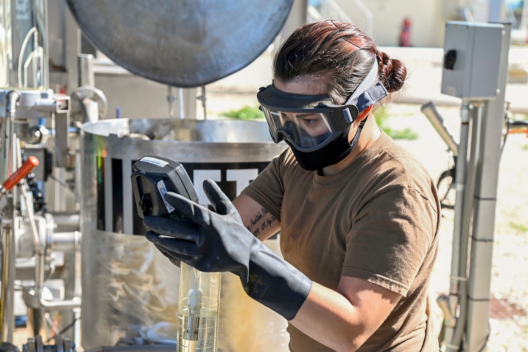 A sailor wearing goggles and black gloves looks at a handheld device while standing amid industrial-type equipment.