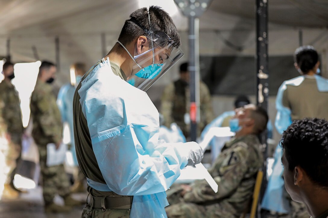 A soldier in a blue gown, mask and face shield works in a room where other soldiers are seated or standing in a line.