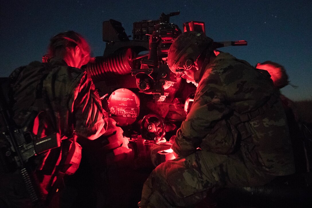 Paratroopers work on a large gun under a red light at night.