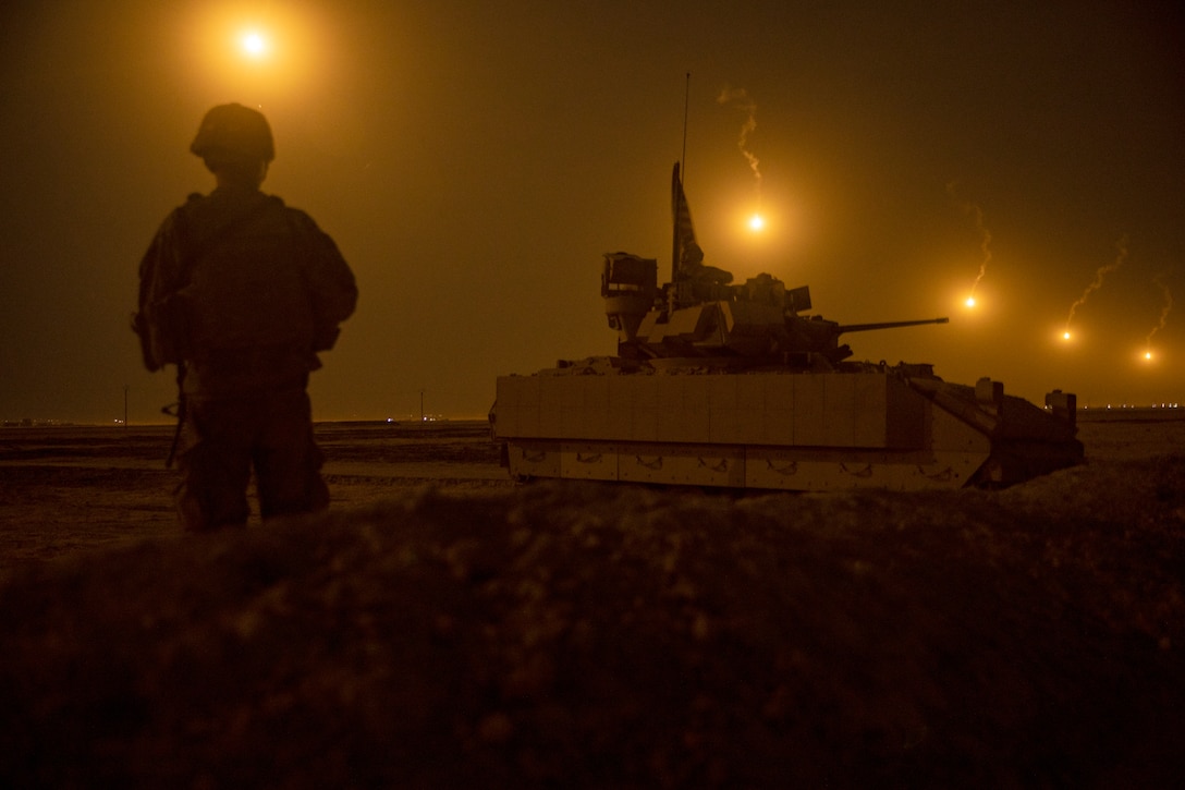 A soldier stands next to large tank at night.