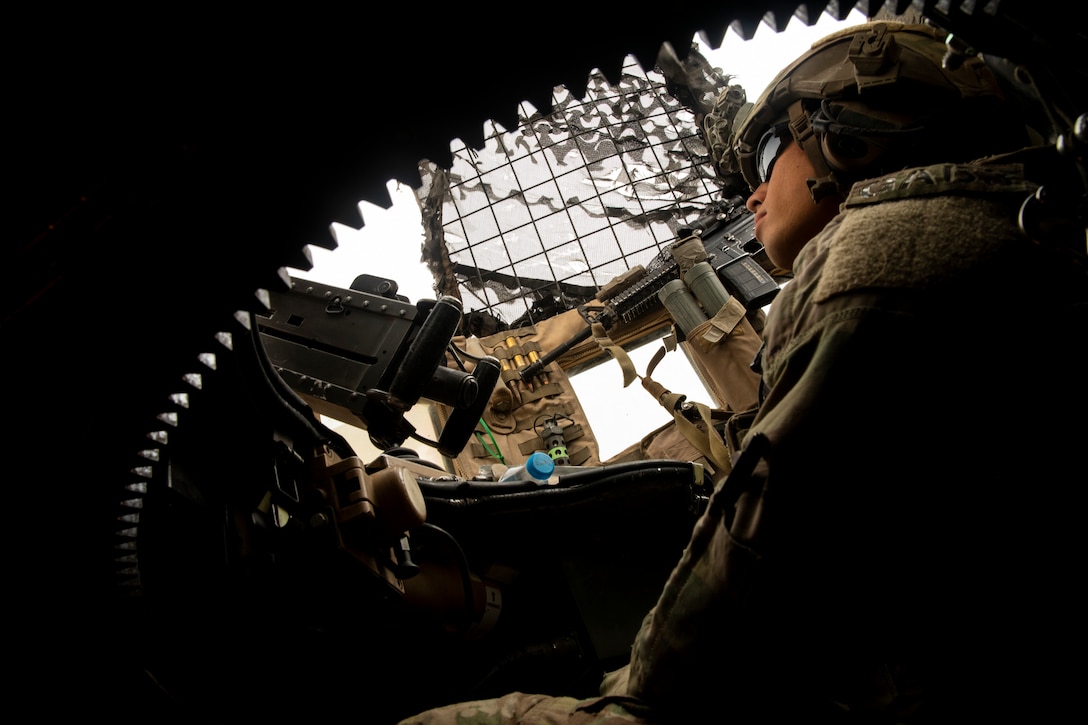 A soldier sits in a military vehicle looking out of a small hatch.