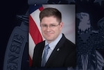 Head and shoulders shot of a white man in a suit sitting in front of the stars and stripes of the US flag.