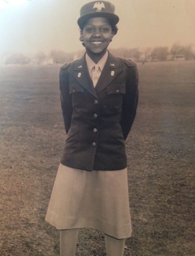 A smiling woman in a ball cap stands with her hands behind her back.