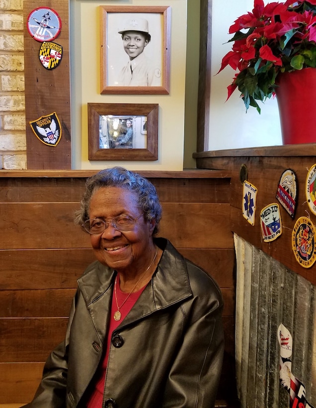 A smiling elderly woman sits in a booth. Photos are on the wall above her.